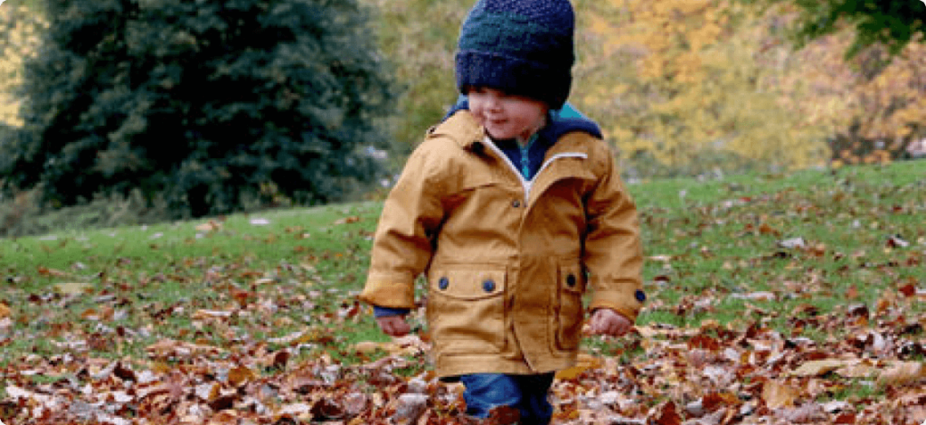 a young boy in a yellow jacket and black hat walking through autumn leaves