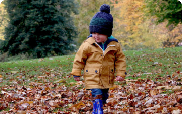 a young boy in a yellow jacket and black hat walking through autumn leaves